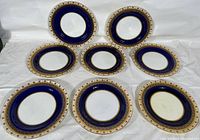 Group of eight antique Minton dinner plates arranged on a white surface showing the front of the plates with dark blue and gold patterned borders.