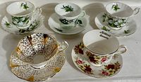 Five vintage fine bone china tea cups and saucers displayed side by side showing varied floral designs, green and pink motifs, and gold accents.