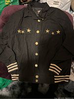Black letterman style jacket with gold color star embellishments on chest and striped cuffs, laid flat showing front button closure.