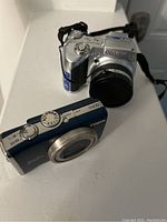 Two digital cameras on a white surface; Canon PowerShot SX200 IS (dark blue and silver) and Olympus SP-510UZ (silver) positioned showing top and front views, demonstrating condition and model details.