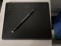 Top-down photo showing the entire Huion graphics tablet with dotted active area and four shortcut buttons on each side. The black Adonit Mini 4 pen is placed diagonally on the tablet.