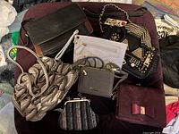 Seven small bags displayed on a surface including a black Alexander Henry leather bag, a studded black handbag, metallic woven bag, fabric coin purse, white wristlet, maroon magnetic bag with bow, and a light gray zipper pouch.