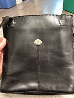 Front view of the black leather crossbody bag showing smooth leather surface and metal logo badge with strap attachment points visible.