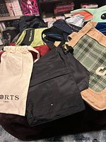 Photo showing various bags including a black Tommy Hilfiger bag, beige canvas tote with green graphic, and others piled on a surface.