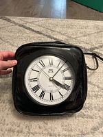 Front view of the square black patent vinyl handbag shaped like a clock with white clock face and black Roman numerals, showing brand Marilyn Brooks and quartz marking.