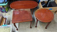 Three wood veneer tables: one oval coffee table, one square side table, one round side table. Slight scratches visible on surfaces.