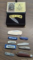 Displaying all eight knives, including the commemorative George Washington folding knife in a presentation box with gold handle, and multiple small pocket knives with Polaroid branding and varied handles.