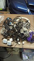 Photo showing a large tangled pile of mostly black and white electronic cables, chargers, and power adapters spread on a wood table.