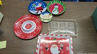 Top view of six holiday-themed serving plates and trays on wooden table, showing colorful festive designs with Santa, snowmen, and snowflakes.