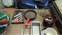 Overview of the kitchen lot on a wooden surface, showing cast iron skillet with red handle, boxed kitchen knives, glass baking dish, metal loaf pan, ceramic crock, bowls, metal stand and kitchen cloths.