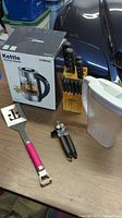 Photo of electric kettle in box, wooden knife block holding knives, metal spatula with pink handle, manual can opener, and a plastic container with lid laid out on table.