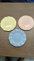 Three scalloped ceramic plates in yellow, pink, and blue on a wood surface.