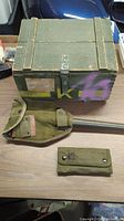 Green wooden ammunition box with visible latch and markings with purple paint over it. Included folding entrenching tool with metal blade and canvas cover, plus a small green canvas pouch.