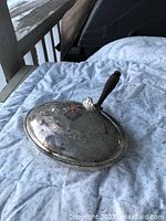 Photo shows an antique silver plated warming dish resting on a cloth, with its lid on. The lid is decorated with ornate engraving and topped with a knob attached to a wooden handle.