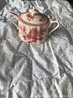 Front angle photo of antique ceramic sugar bowl with red pattern and lid on fabric background.
