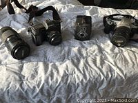 Full view of four main camera items on bed including two SLR cameras, one box camera, and one telephoto lens.