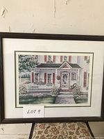 Framed and signed print by Janice Tanton showing a house scene, surrounded by a dark brown frame with a white mat.