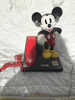 Photo of a vintage Mickey Mouse novelty telephone with a red handset on a black base. Mickey Mouse figure is standing upright on the right side of the base.