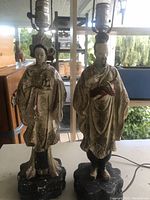 Pair of Asian figurine lamps standing side by side on a table under a covered porch area with background furniture and greenery.