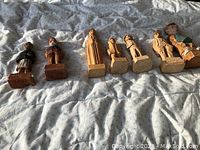 Seven hand carved wooden figurines arranged in a row on fabric background. Varied designs including painted and natural wood tone pieces.
