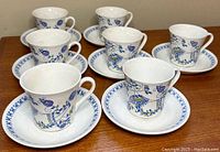 Seven white ceramic cups and saucers with blue floral and character designs, arranged on wooden surface, showing full set.