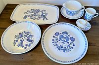 Overall view of the Figgjo Norway vintage dish set showing rectangular platter, shallow bowl, dinner plate, large cup with saucer, egg cup, and small handled cup. All pieces have blue and purple floral and human figure designs on white ceramic.