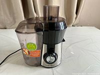Front right angled view of the juicer showing the main unit with control knob, power cable, and juice container attached.