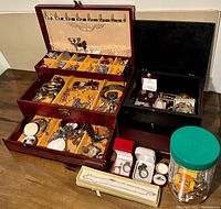 Wide front and top view of large wooden jewelry box opened to reveal top, middle and bottom compartments filled with jewelry plus a smaller black box and a plastic jar with more jewelry placed adjacent on table.