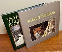 Two hardcover Robert Bateman art books, side by side on a wooden surface, showing front covers with dust jackets. The Art of Robert Bateman has a gray cover with a fox image; The World of Robert Bateman has a green cover with a nature scene.