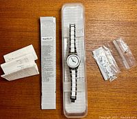 Full lot showing white and silver men's Swatch wristwatch in transparent Swatch-branded plastic box, original papers, and additional watch links.