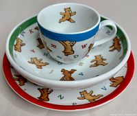 3-piece baby set stacked: mug, bowl, plate showing teddy bear and alphabet design with colored rims