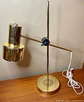 Full lamp profile showing brass cantilever adjustable arm, round base, vertical pole, and white cord coiled on table.