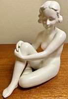 Front angle of white porcelain nude figurine seated with arms wrapped around knee on wood surface.