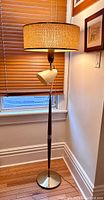 Full view of the floor lamp with woven beige fabric shade, teak wood central column, and metal base
