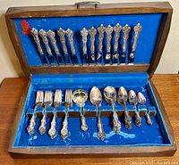 Full view of open wooden chest with blue satin/velvet interior containing the silverplate flatware laid out, showing knives, forks, spoons arranged in slots.