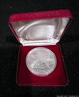 The coin inside a red velvet presentation box with white satin lining, showing the coin encased in plastic.