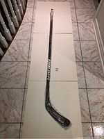 Full length image of the Bauer Vapor hockey stick, showing the stick fully wrapped in original plastic packaging on a tiled floor.