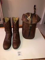 Pair of brown leather cowboy boots and a brown leather saddle bag displayed upright on a white surface showing overall condition and wear.