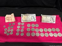 Complete view of Canadian coins and bills spread on a red cloth including dollar coins, fifty cent pieces, quarters, dimes, one and two dollar bills.