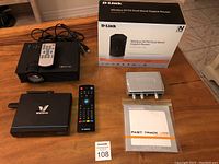 All items displayed on a wooden table including the Blitzwolf projector, D-Link router with box, M-Audio audio interface with manual, VMedia TV box and remote controls.