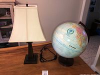 Photo showing Pottery Barn table lamp with bronze metal base and cream lampshade next to a Globemaster globe on wooden base on table.