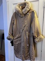Front view of beige Linda Lundstrom hooded coat with fur trim on hood and cuffs, showing asymmetrical closure and large pockets.
