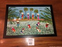 Framed oil painting showing a vibrant Brazilian cotton harvest scene with people harvesting cotton in a field under palm trees, with a black frame and wood surface background.