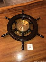 Oak ships wheel clock on wooden surface, clock face showing time with black numbers and red second hand, wooden spokes visible.