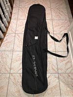 Black Dakine snowboard bag lying flat on tiled floor, featuring carrying strap and side pockets.