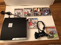 Photo showing Sony PS3 slim console with power cord, one black DualShock 3 controller with USB charging cable, and six PS3 game cases lined at back.