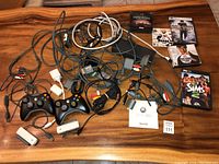 Overview of the lot including 2 Xbox 360 wired controllers, various cables with AV connectors and adapters, and multiple PC game cases and discs including The Sims 2.