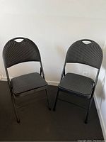 Two folding chairs with padded seats and backs, dark textured fabric upholstery, and black metal frames shown side by side against a white wall.