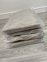 Stack of four beige bath mats, each individually packaged in plastic