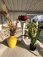 Photo showing three floral arrangements: yellow vase with dried florals, white woven pedestal planter with red flowers, and black pot with green and yellow flowers.